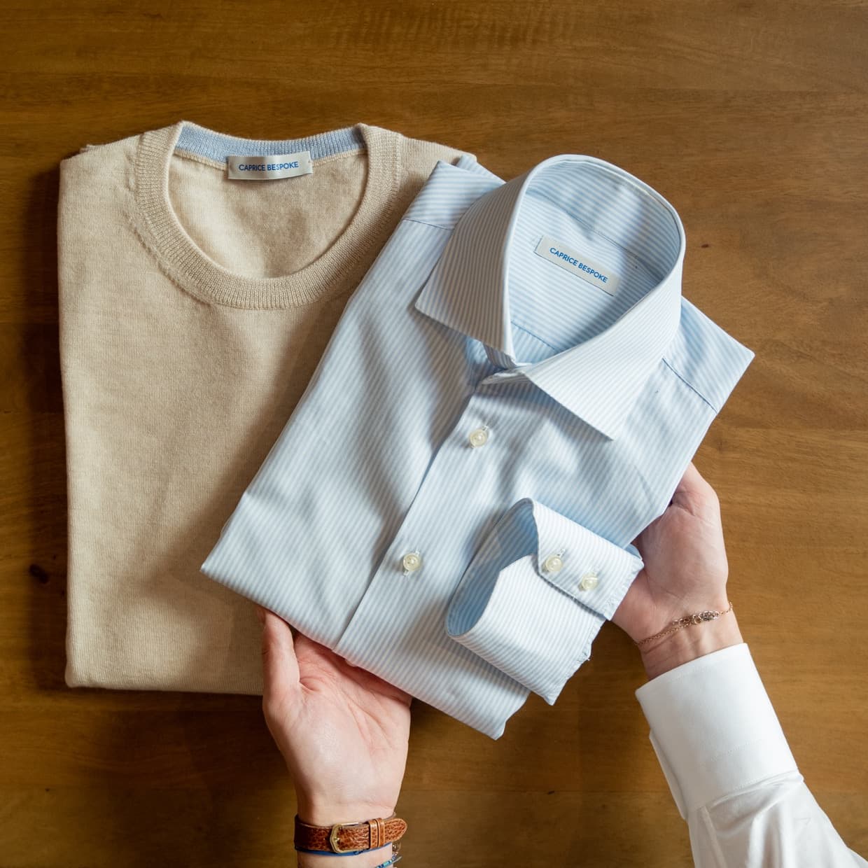 A folded knit and a tailored shirt with a Caprice Bespoke label, held in hand on a wooden table