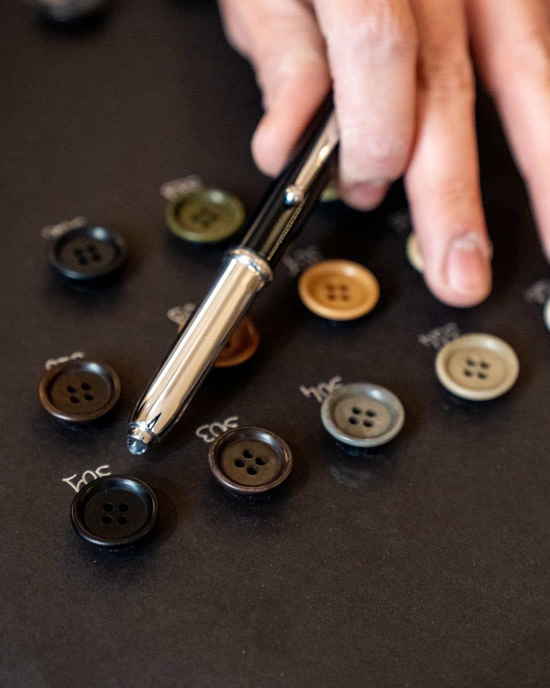 A hand selecting horn buttons from a numbered tray while a garment specification is finalised